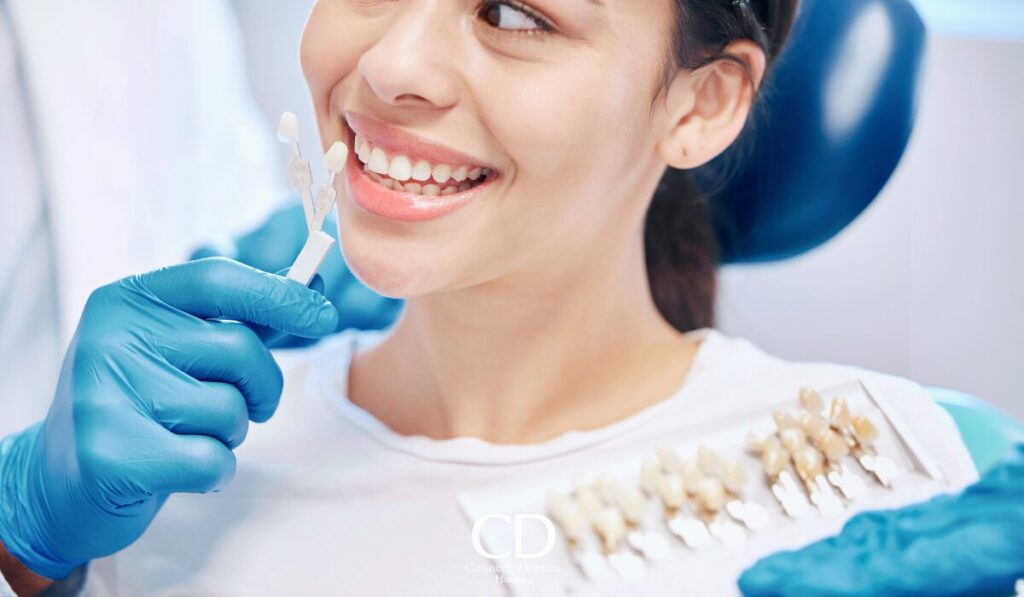 Dentist wearing blue gloves holds shade guide near smiling patient's teeth for color matching.